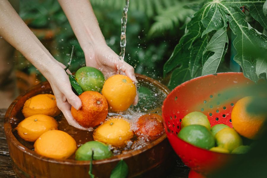 Manos lavando naranjas y limas frescas en un cuenco de madera junto a hojas verdes
